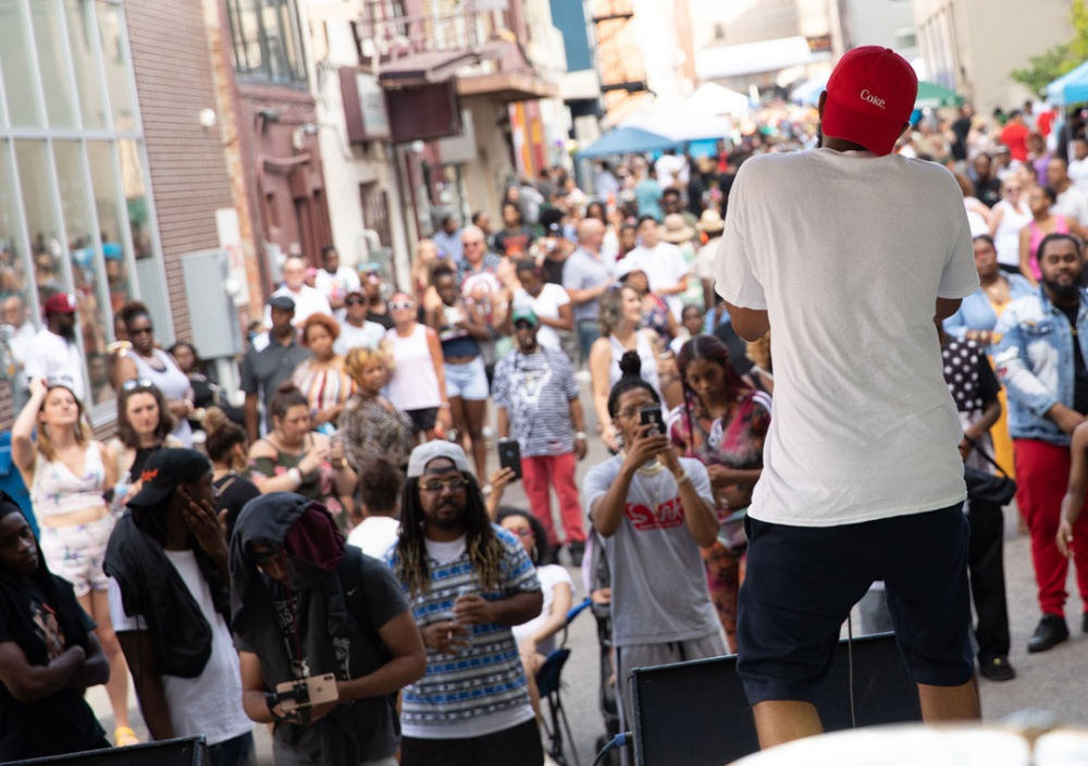 A performer sings to an audience from the stage set up in one of Flint's alleys, called Buckham. His back is to the camera but the audience in front of the stage is facing the camera. Many people are recording and taking photos of the artist with their cell phones and cameras.