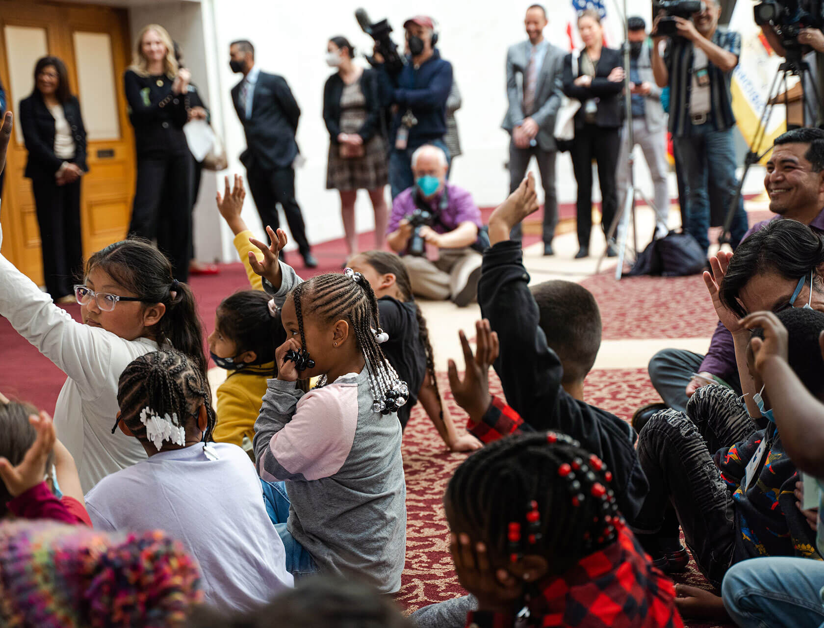 A group of children seated on the floor raise their hands during an event.