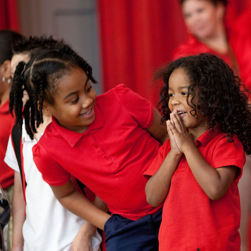 Smiling young girls are on a stage. One looks back at the other, who looks into the distance.