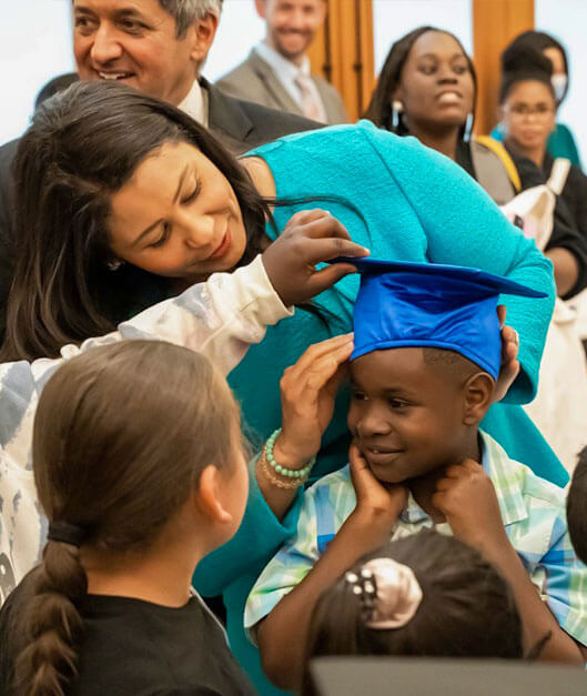 A woman places mortarboard on child’s head, surrounded by other children.