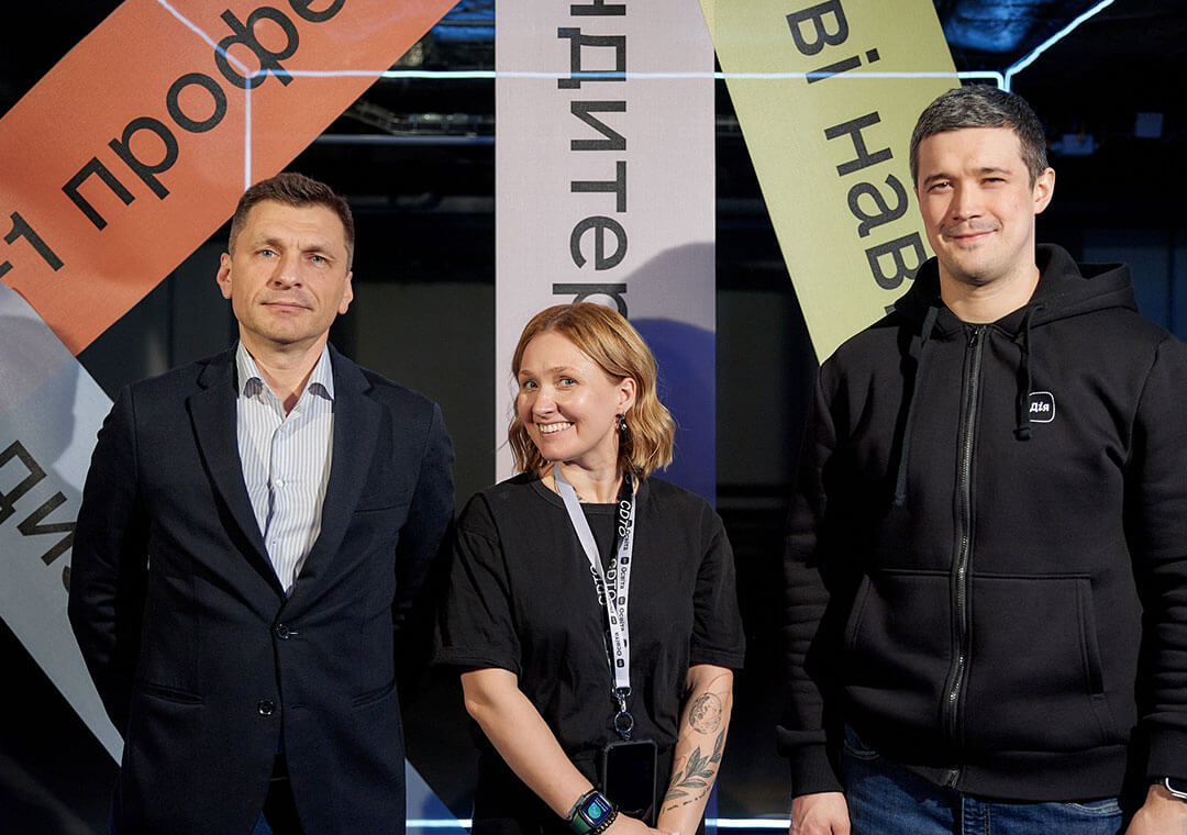 Three people pose in front of colorful banners with Ukrainian writing.