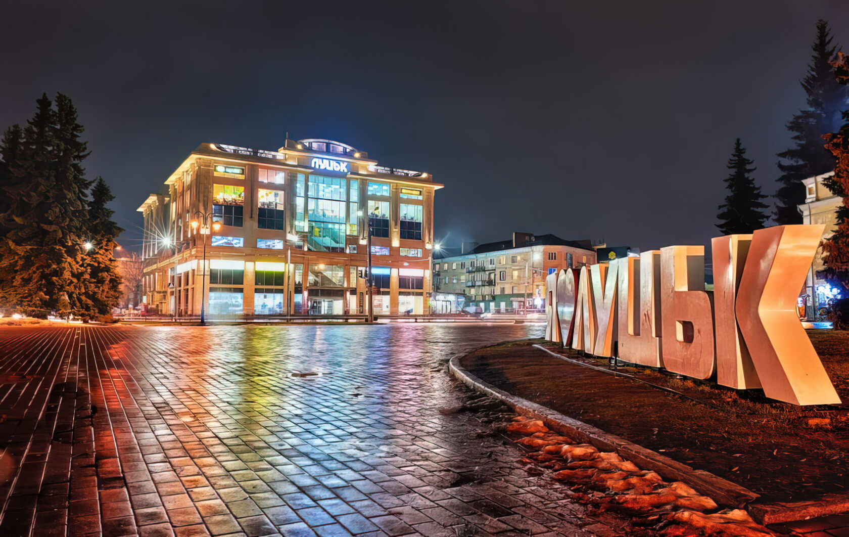 A wet street scene shows a brightly lit shopping center and a large sign made of three dimensional letters that says I "heart" Lutsk. The paving bricks reflect the lights.
