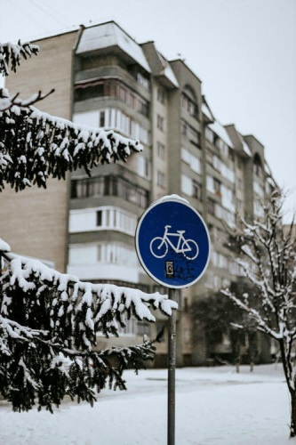 A snow-covered scene shows a road sign for a bike with trees and a building in the background.