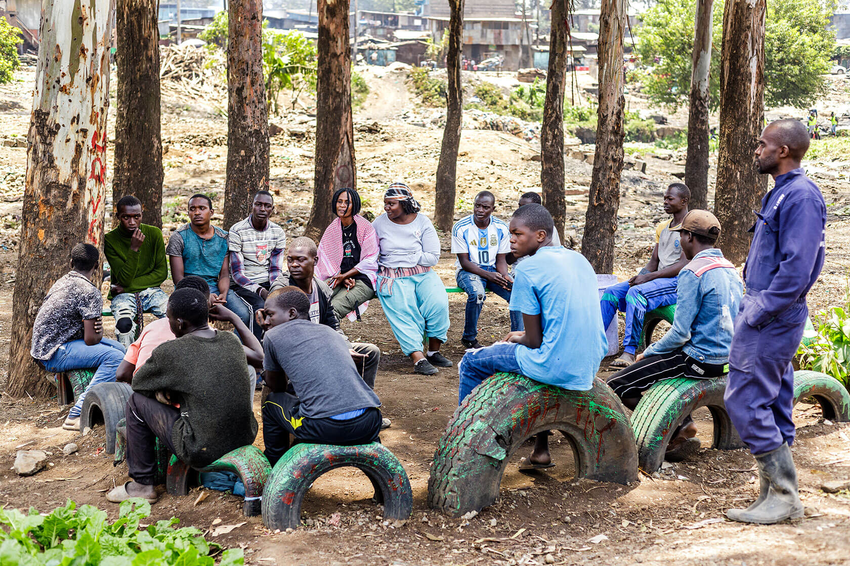 A group of people sit on tires that have been set into the ground to make an informal seating circle. A row of trees and housing are in the background.