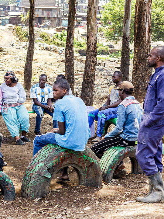 A group of people sit on tires that have been set into the ground to make an informal seating circle. A row of trees and housing are in the background.