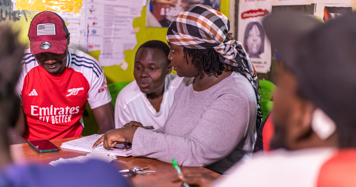 A woman points at a notebook while talking to several others seated at a table. Posters cover the wall behind them.