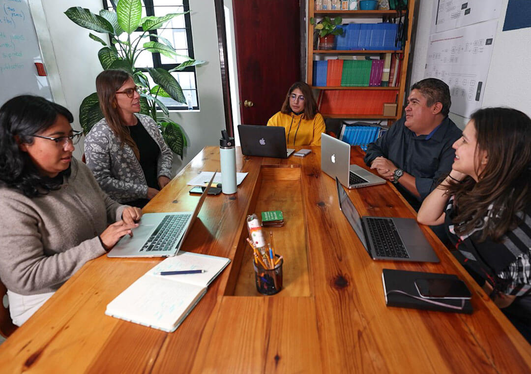 A group of five sit at a wood table in an office space, most with laptops out, having a discussion.