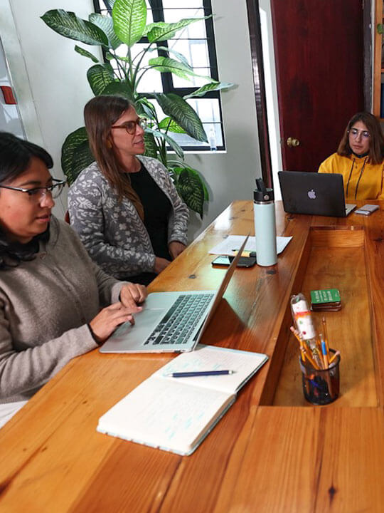 A group of five sit at a wood table in an office space, most with laptops out, having a discussion.
