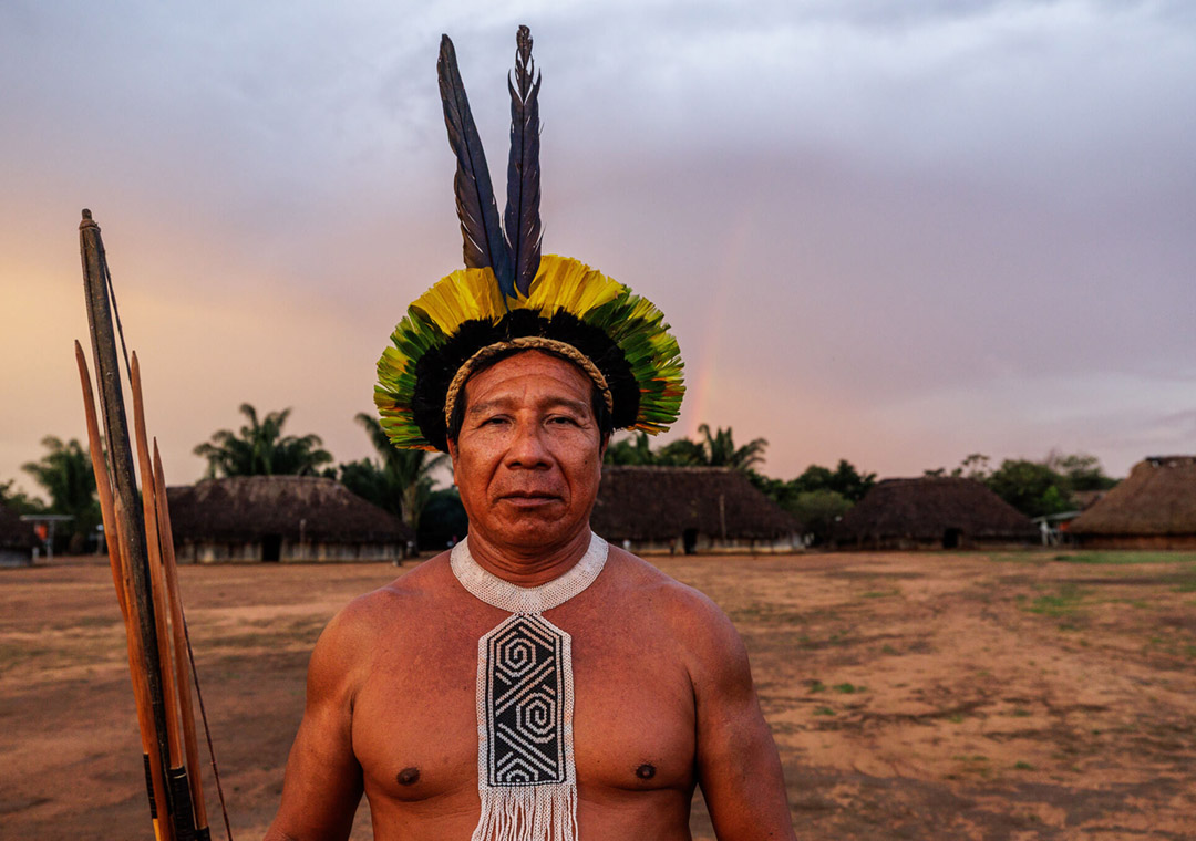 A man who is a leader in the Khikatxi village stands in the center of the village round wearing traditional garb consisting of a head piece while holding a bow and arrows in his right hand as he looks at the camera.