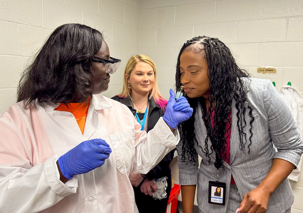 A woman leans over to smell an essential oil bottle held by a student wearing safety glasses, neoprene gloves and a lab coat. Another woman watches.