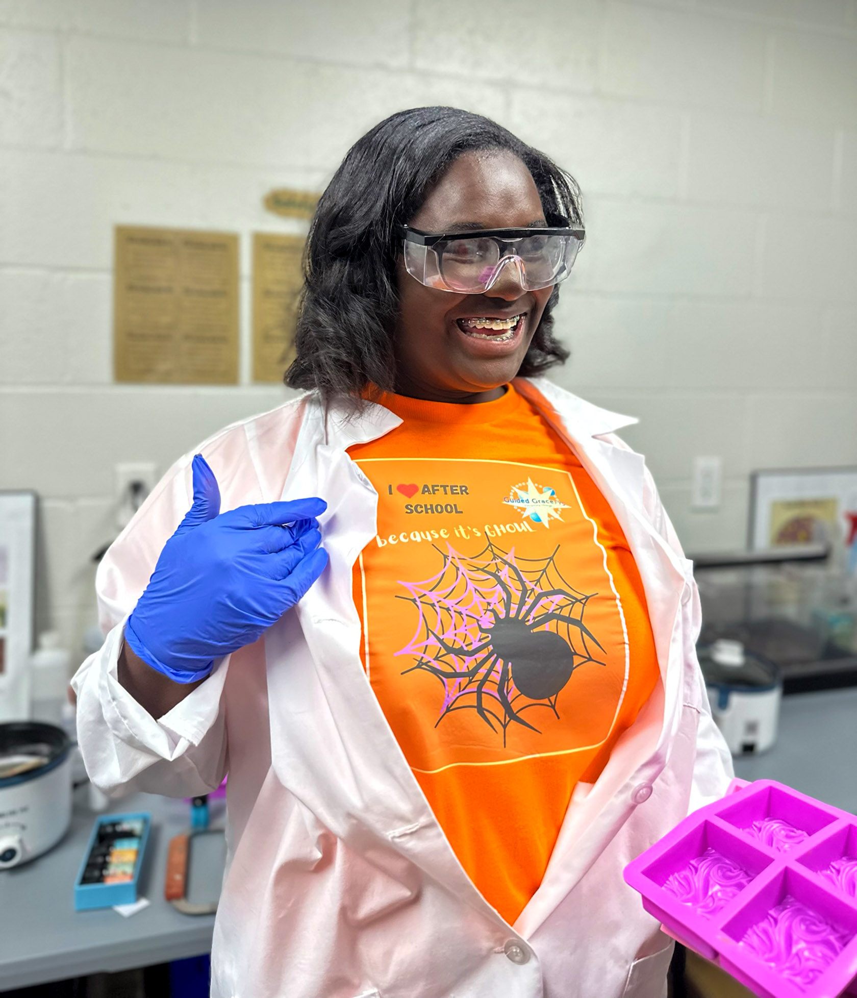 A student wearing safety glasses and neoprene gloves showcases her Halloween-themed “I heart Afterschool” T-shirt design while holding a soap mold.