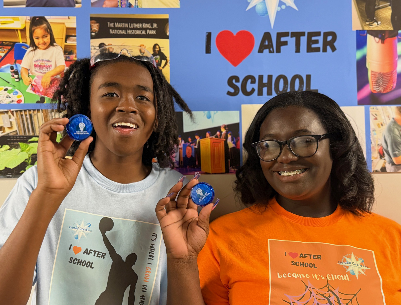 Two smiling students proudly display their Lights On Afterschool campaign buttons, while standing in front of an afterschool themed poster.