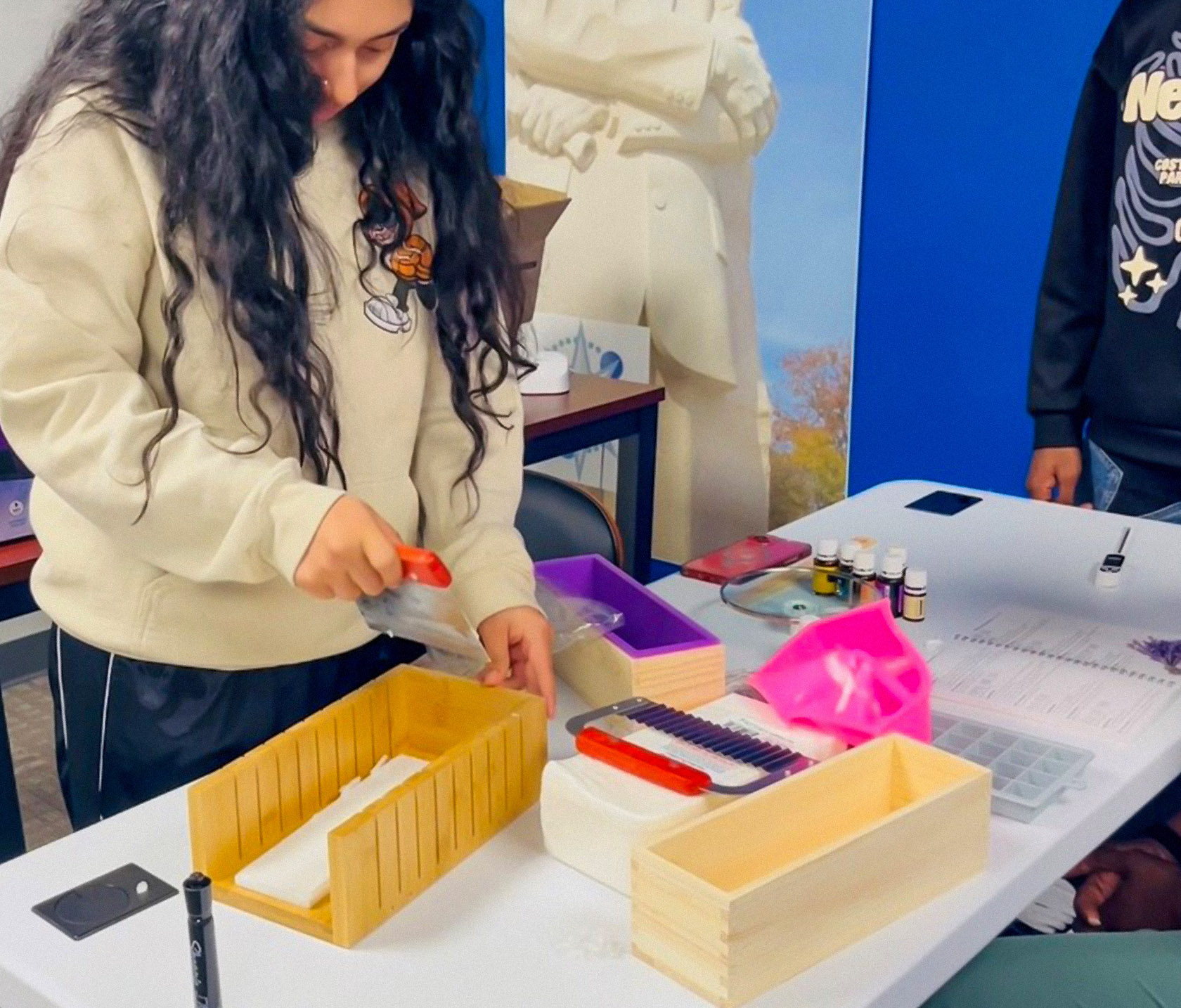 A student practices using a soap cutter while standing at a table with soap molds and essential oils.