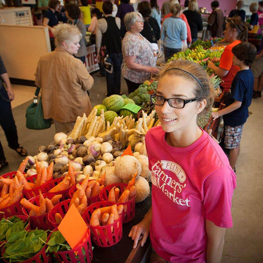 A young woman looks up from a produce stand selling fruit and vegetables. Other people are on both sides of the stand in the background.