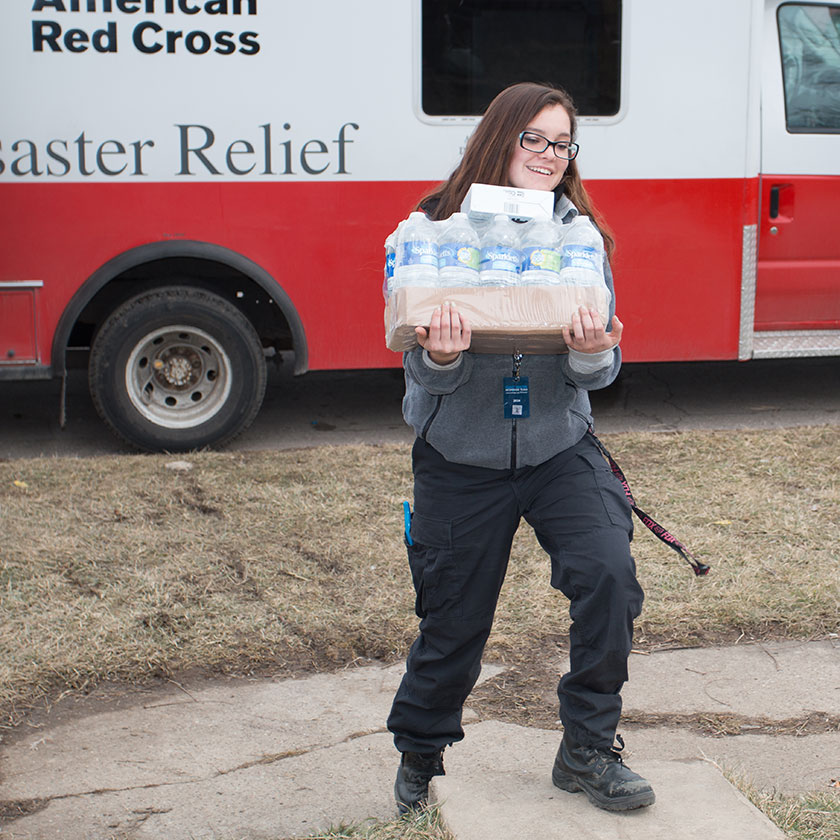 A smiling woman carries a flat of water bottles from an American Red Criss Disaster Relief truck.