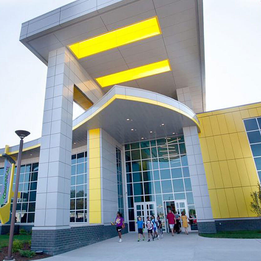 A group walks out the front door of the Flint Cultural Center Academy. The building is modern with yellow accents.