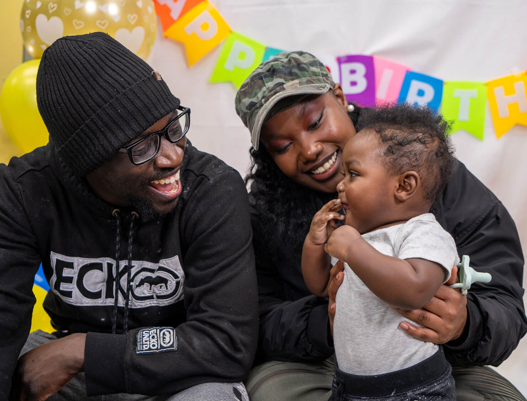 A photo of a young man and woman smiling at their infant son. Behind them are colorful birthday banners and balloons.
