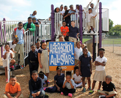 A group of children pose on a playground with a sign that says, “After-School All-Stars.”