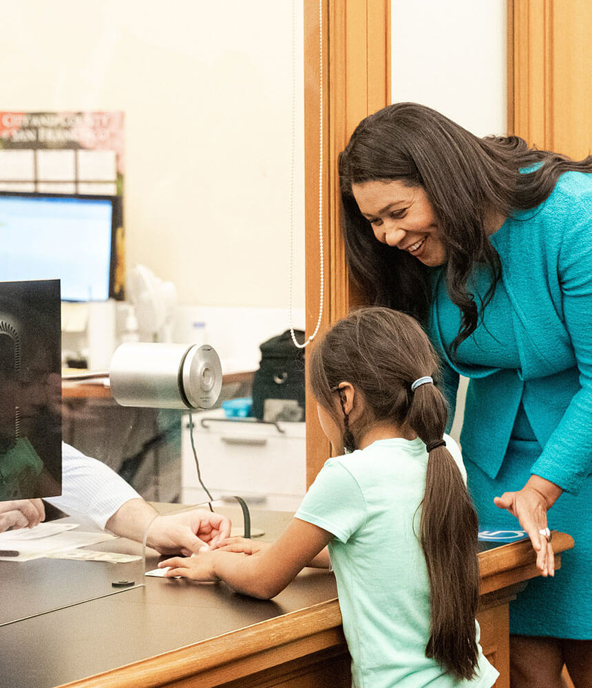 A smiling woman watches over the shoulder of a young girl, who makes a deposit with a bank teller.