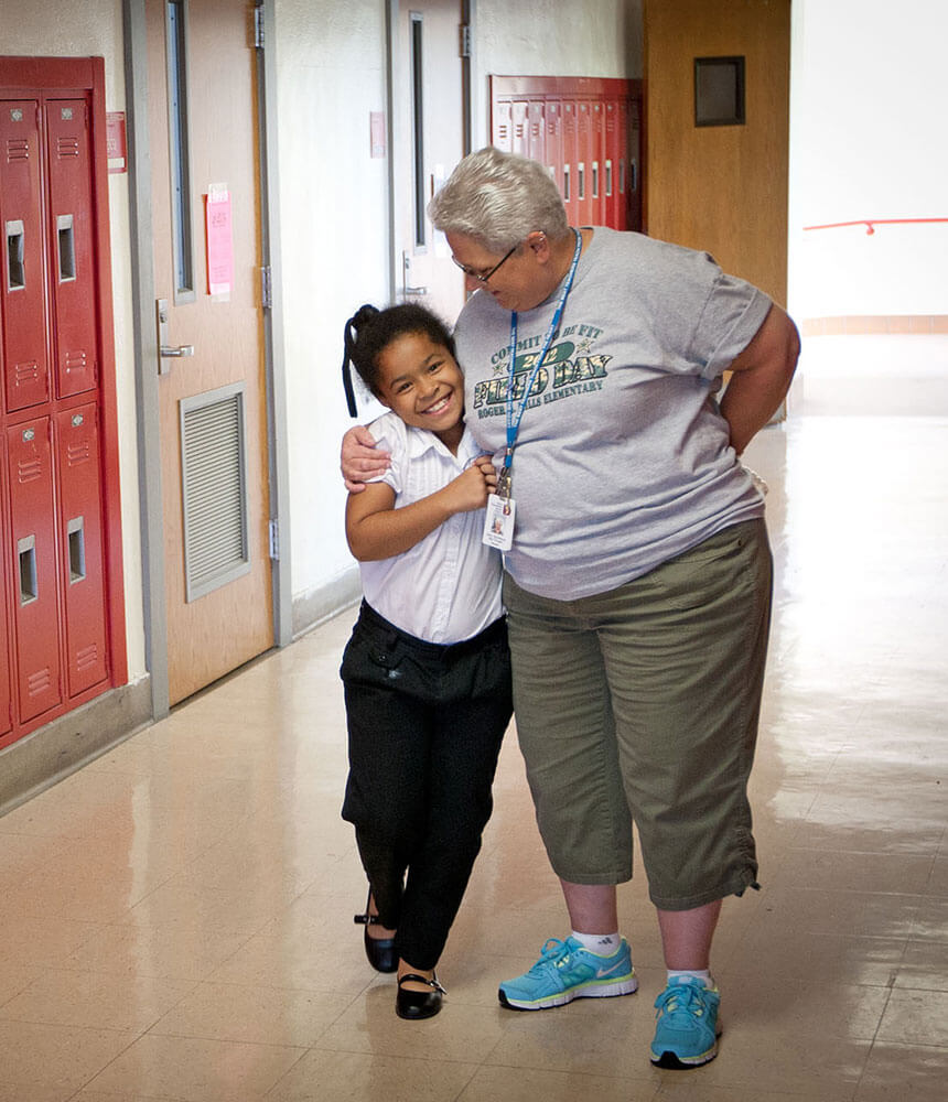 A woman hugs a smiling young girl from the side as they walk past a row of red lockers in a school hallway.