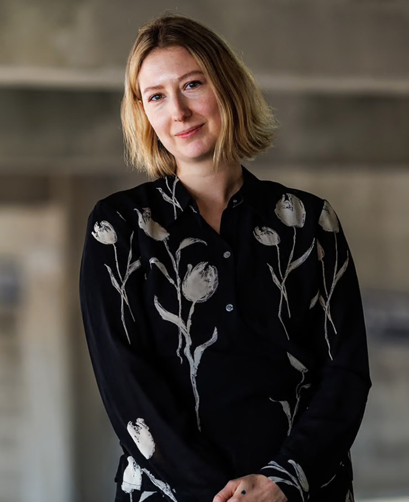 A woman with a blonde bob hair in her 30s wears a black blouse with tulips on it smiles at the camera.