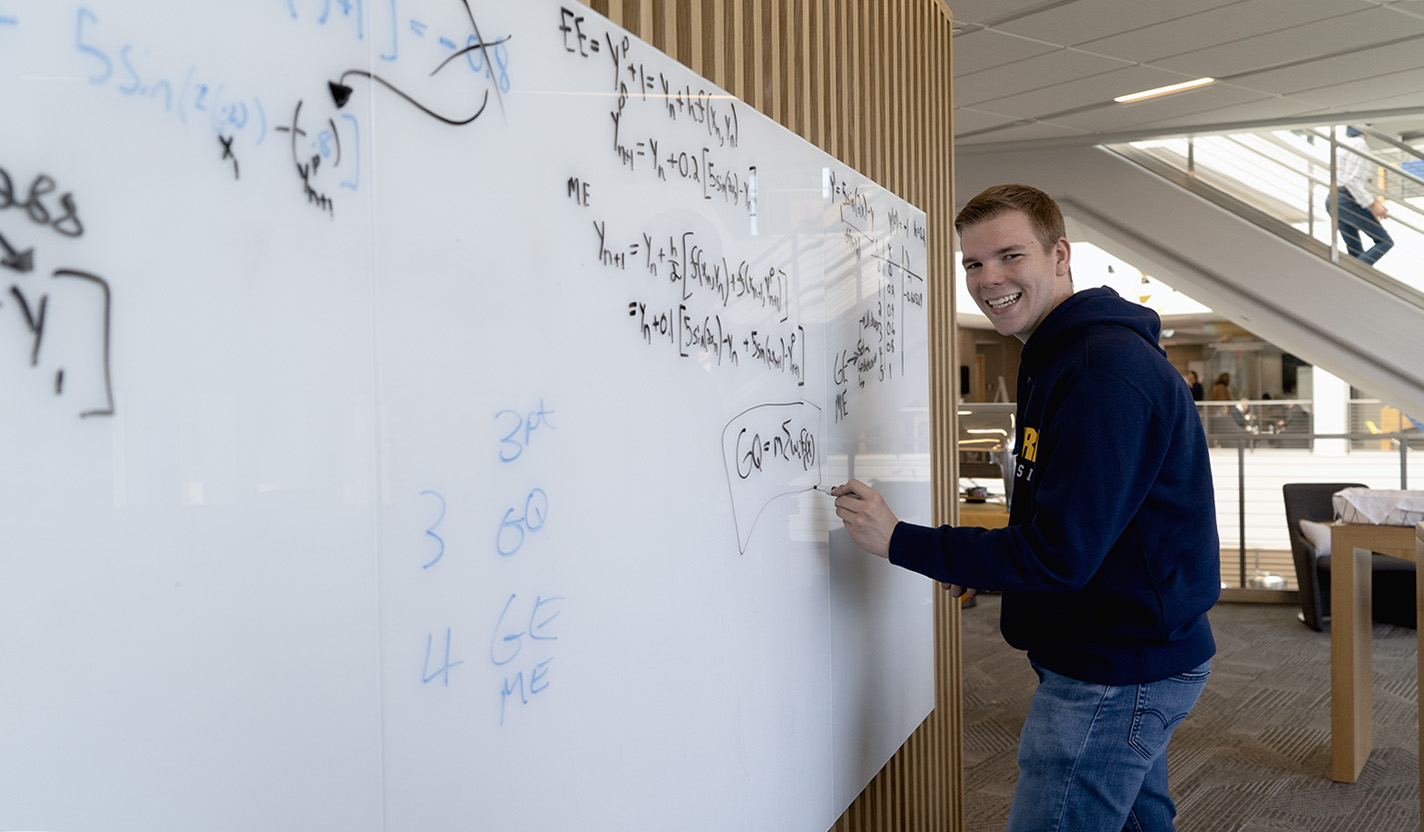 A male student smiles at the camera as he writes on a whiteboard covered with math equations.