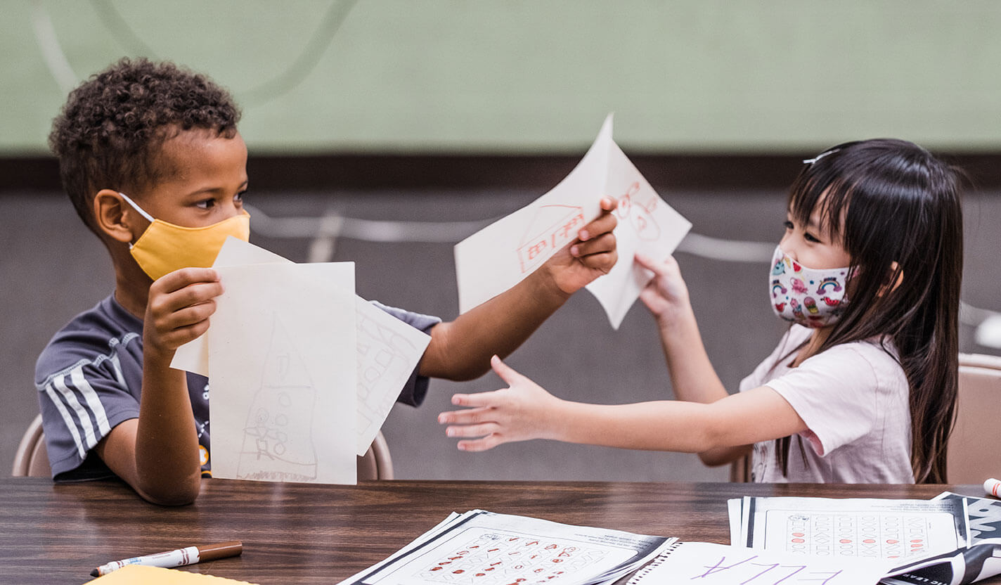 Two small children in masks exchange drawings.