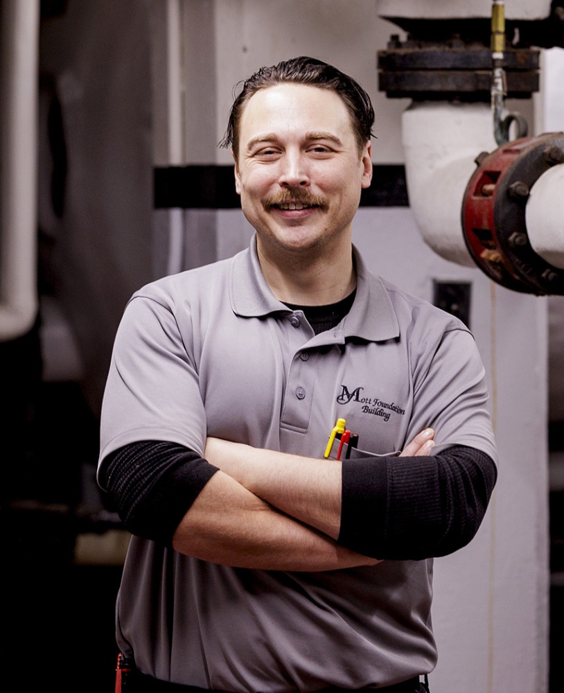 A man poses with arms crossed near pipes industrial pipes in the basement of a 16 floor building.