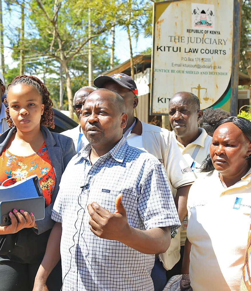 A group stand with a man speaking to someone off-camera. A "Republic of Kenya, the Judiciary, Kitui Law Courts" sign is in the background.
