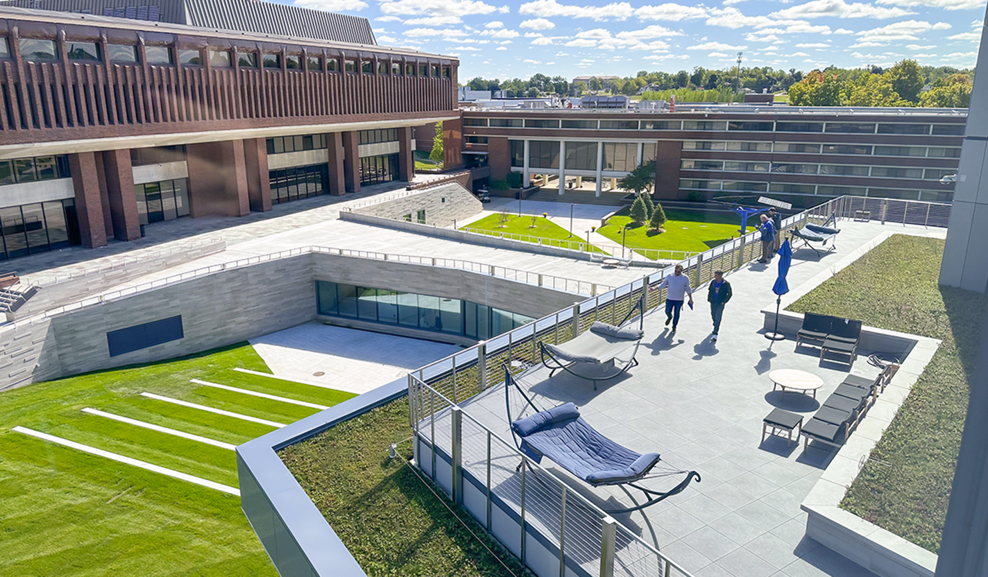 Two people are walking outside on a terrace with hammocks and other chairs around them. They overlook campus buildings and a grassy courtyard.
