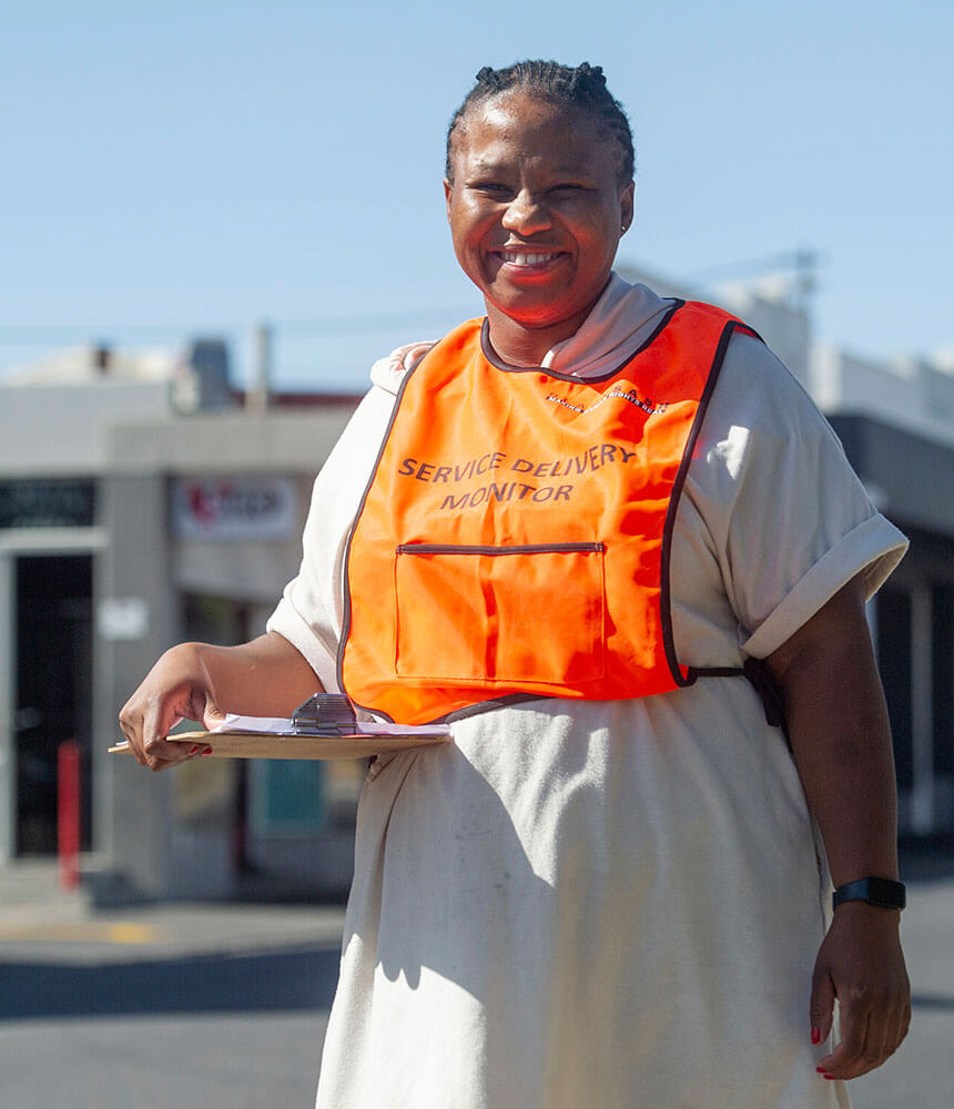 A smiling woman holding a clipboard wears a bright orange "service delivery monitor" vest.