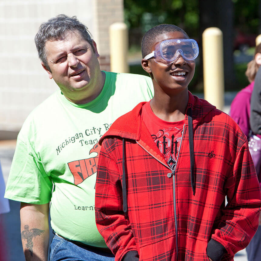 A man looks over the shoulder of a smiling teen boy wearing safety goggles as they watch a rocket launch off screen.
