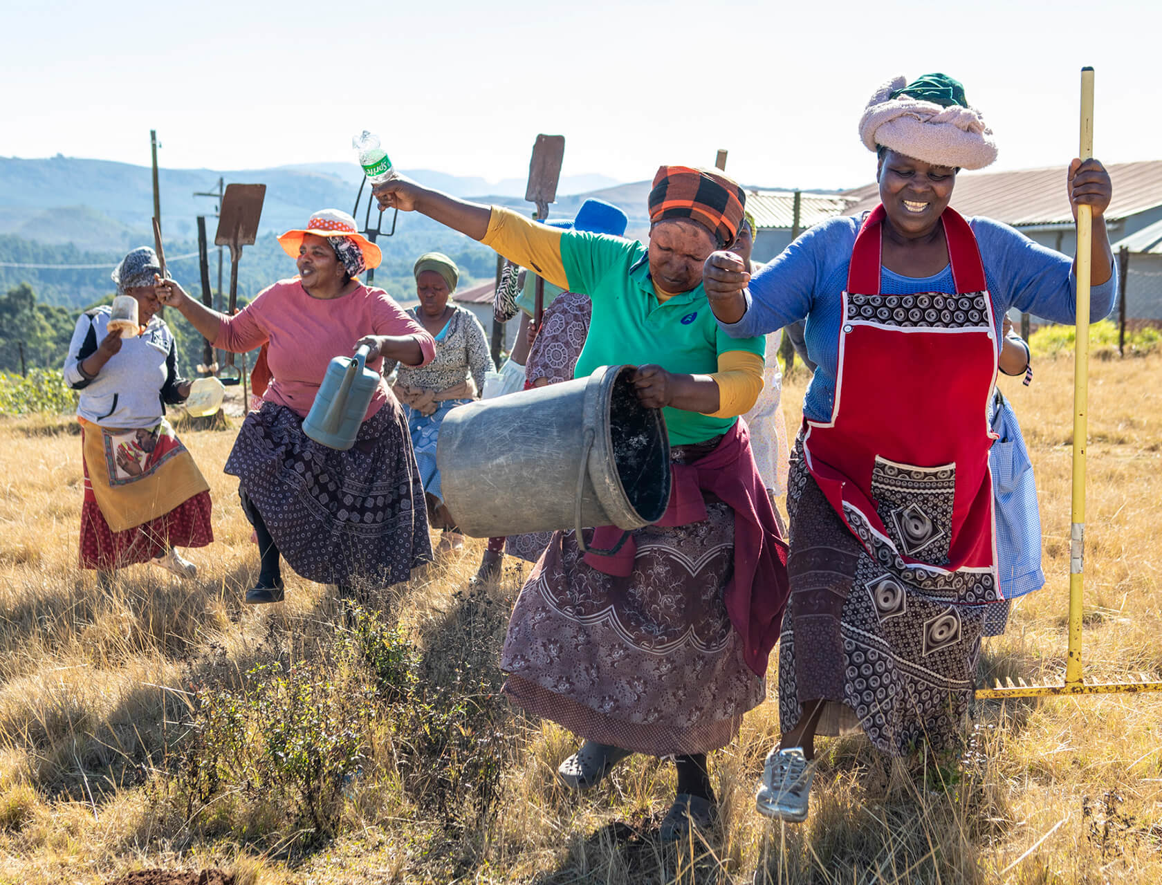 A group of people, mostly women, work together outdoors in a field. They carry buckets, bottles, and farming tools, wearing aprons and head coverings, with hills and buildings in the background.