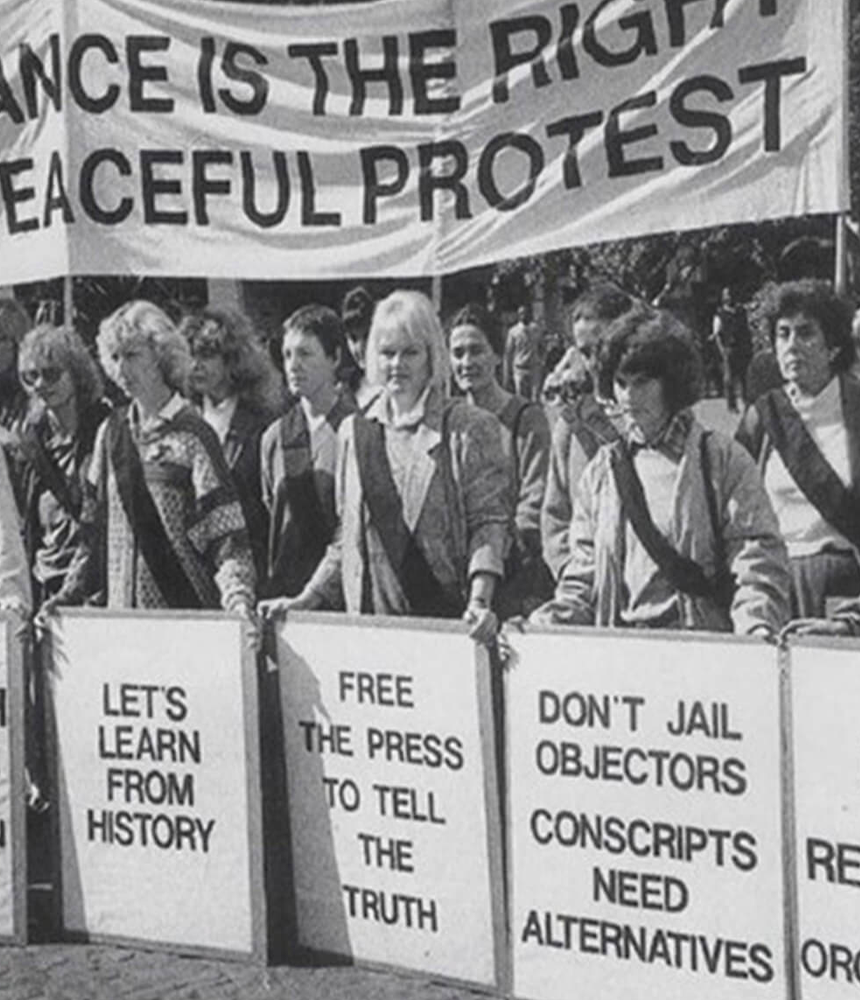 A black-and-white photo of a protest. A line of people stand holding large signs with messages such as “Abolish apartheid legislation,” “Let’s learn from history,” and “Free the press to tell the truth.” A large banner above reads “Defiance is the right to peaceful protest.”