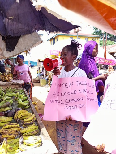 A person holds a megaphone and a pink sign that reads “Women deserve a second chance in the criminal justice system” at a busy outdoor market. Another person stands nearby, and bananas are displayed on a table.