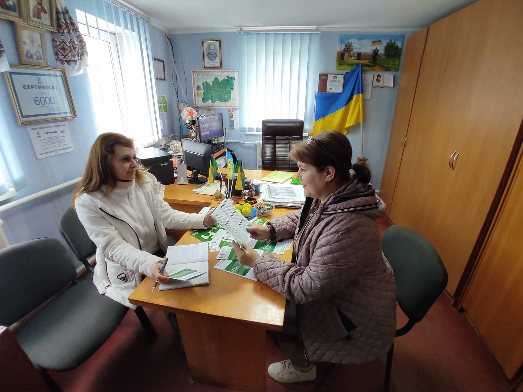 Two people are seated at a desk in an office, exchanging documents. The room contains a Ukrainian flag, framed certificates, and various office supplies. Photo: Courtesy of Ukrainian Legal Aid Foundation