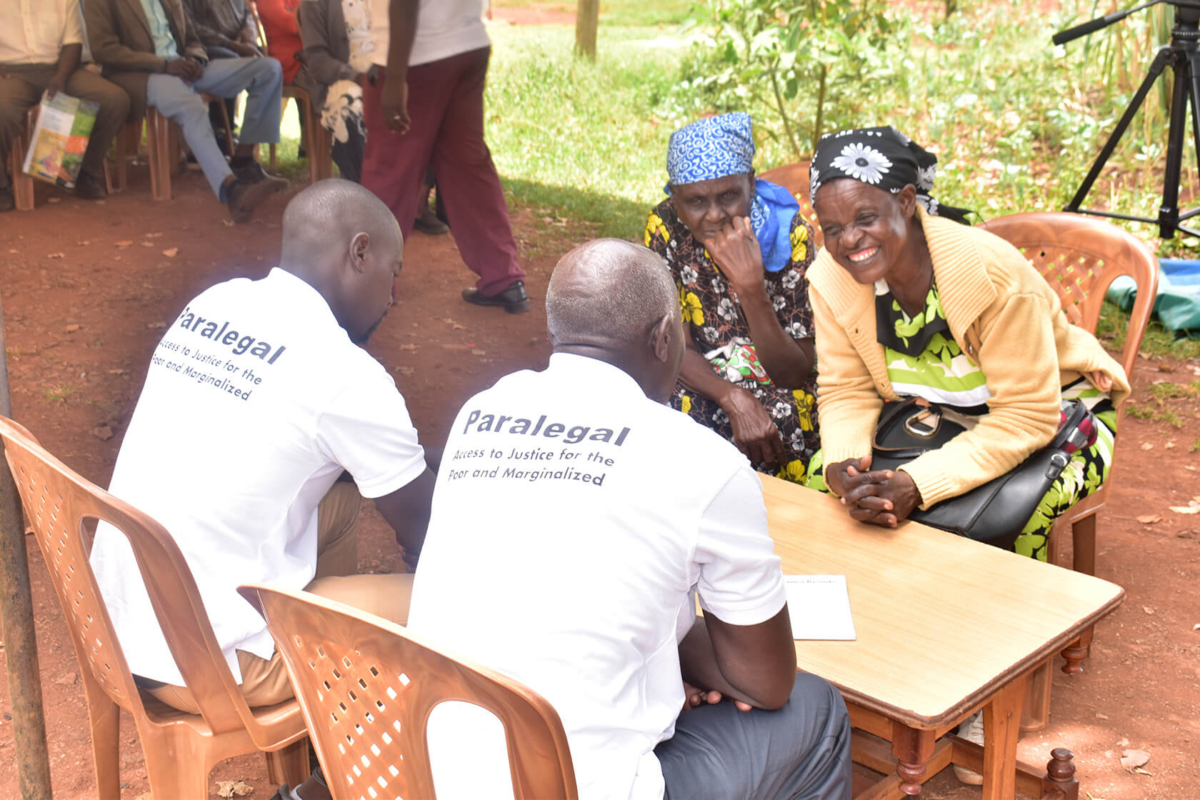 Two people wearing white shirts labeled “Paralegal” sit at a small table with two others, having a discussion in an outdoor setting with more people and greenery in the background.