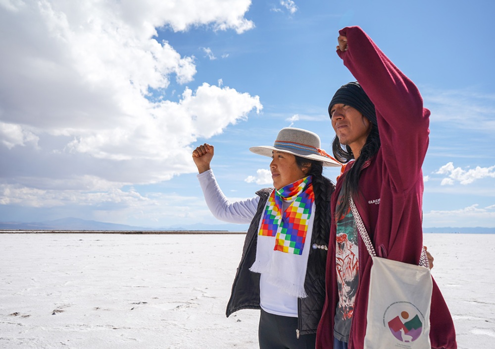 Two people stand together on a vast, bright salt flat under a partly cloudy sky, each raising a fist in the air. One person wears a wide-brimmed hat, a colorful woven scarf, and a dark vest, while the other wears a maroon jacket and carries a shoulder bag with a circular multicolor emblem. Mountains are visible in the distance beneath the expansive sky.