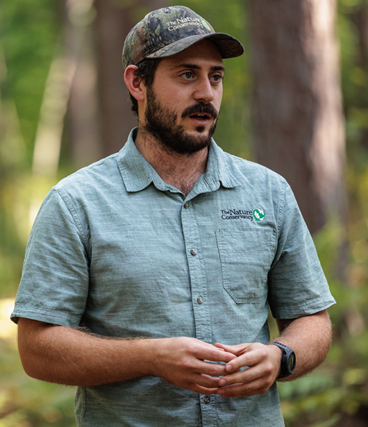 A person wearing a gray short-sleeve button-up shirt with The Nature Conservancy logo and a camouflage cap stands in a sunlit forest, with tall trees and green foliage in the background. Their hands are loosely clasped in front of them.