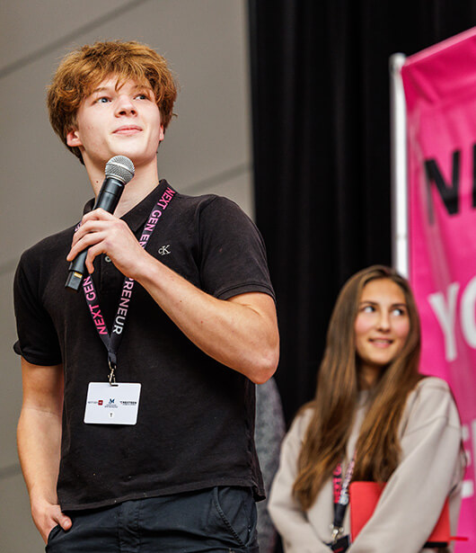 Young man in a black polo speaks into a microphone, standing in front of two other young people.