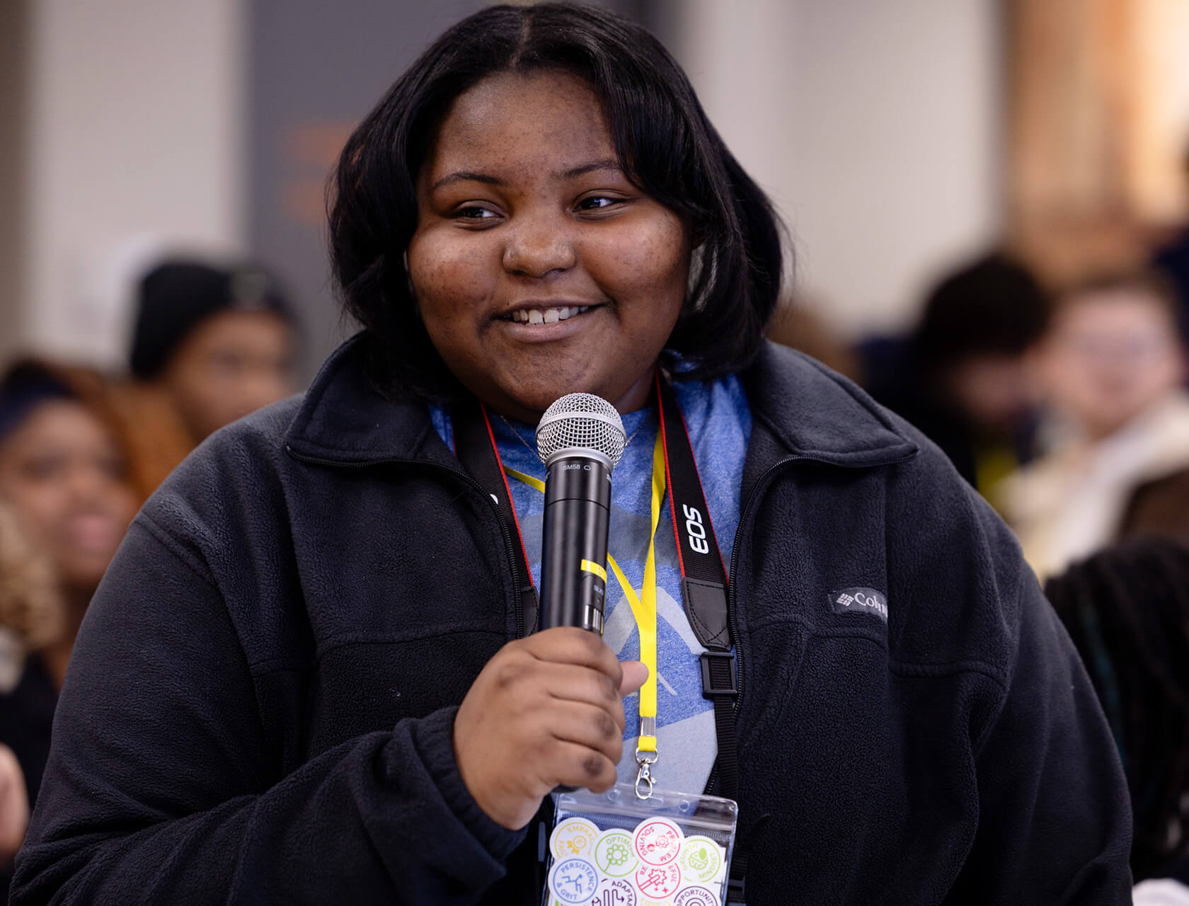 A young woman in a black fleece smiles, speaking into a microphone.
