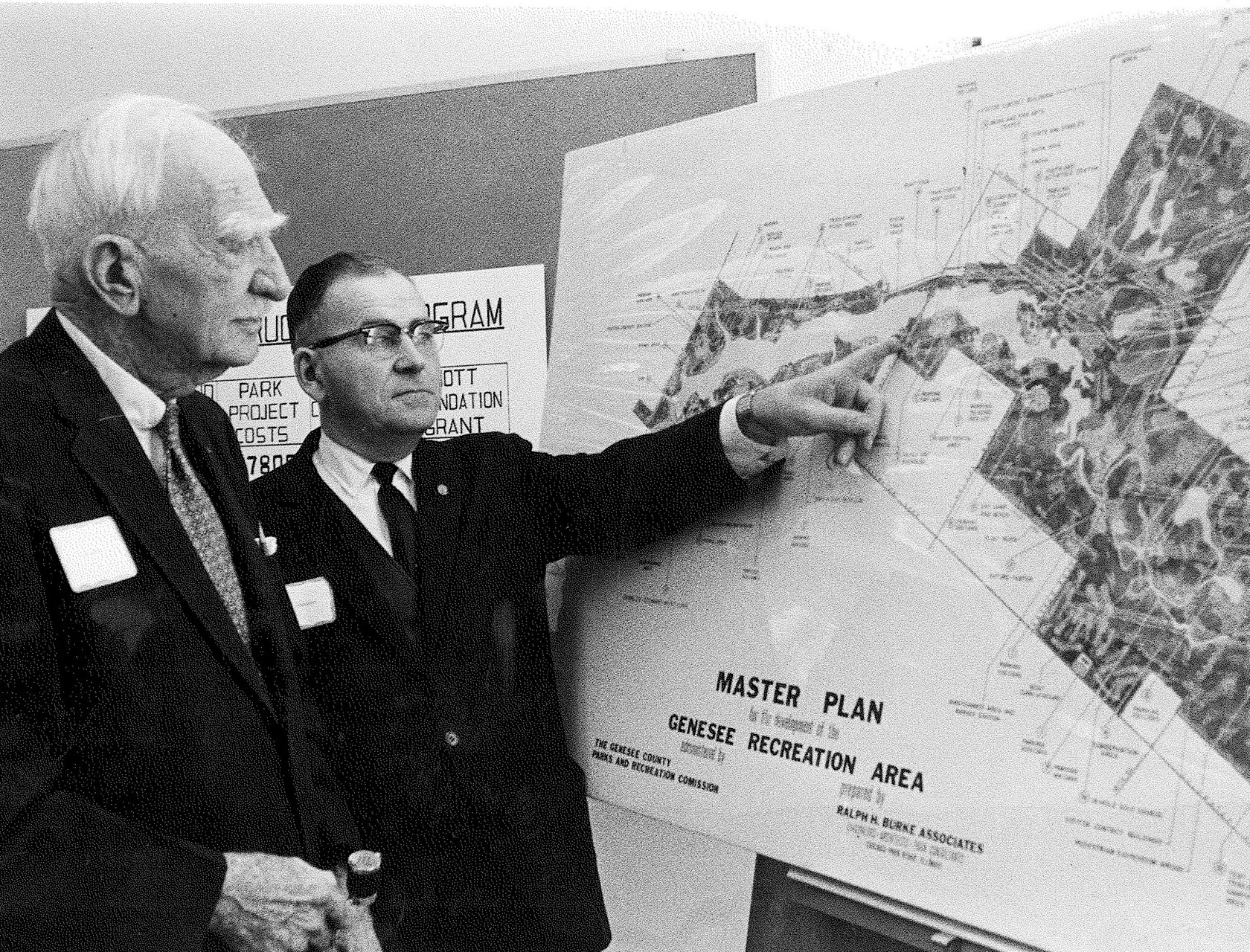 A black and white photo shows two men looking at a map with one man pointing to the map and a title in the bottom left corner reading “Master Plan Genesee Recreation Area.”