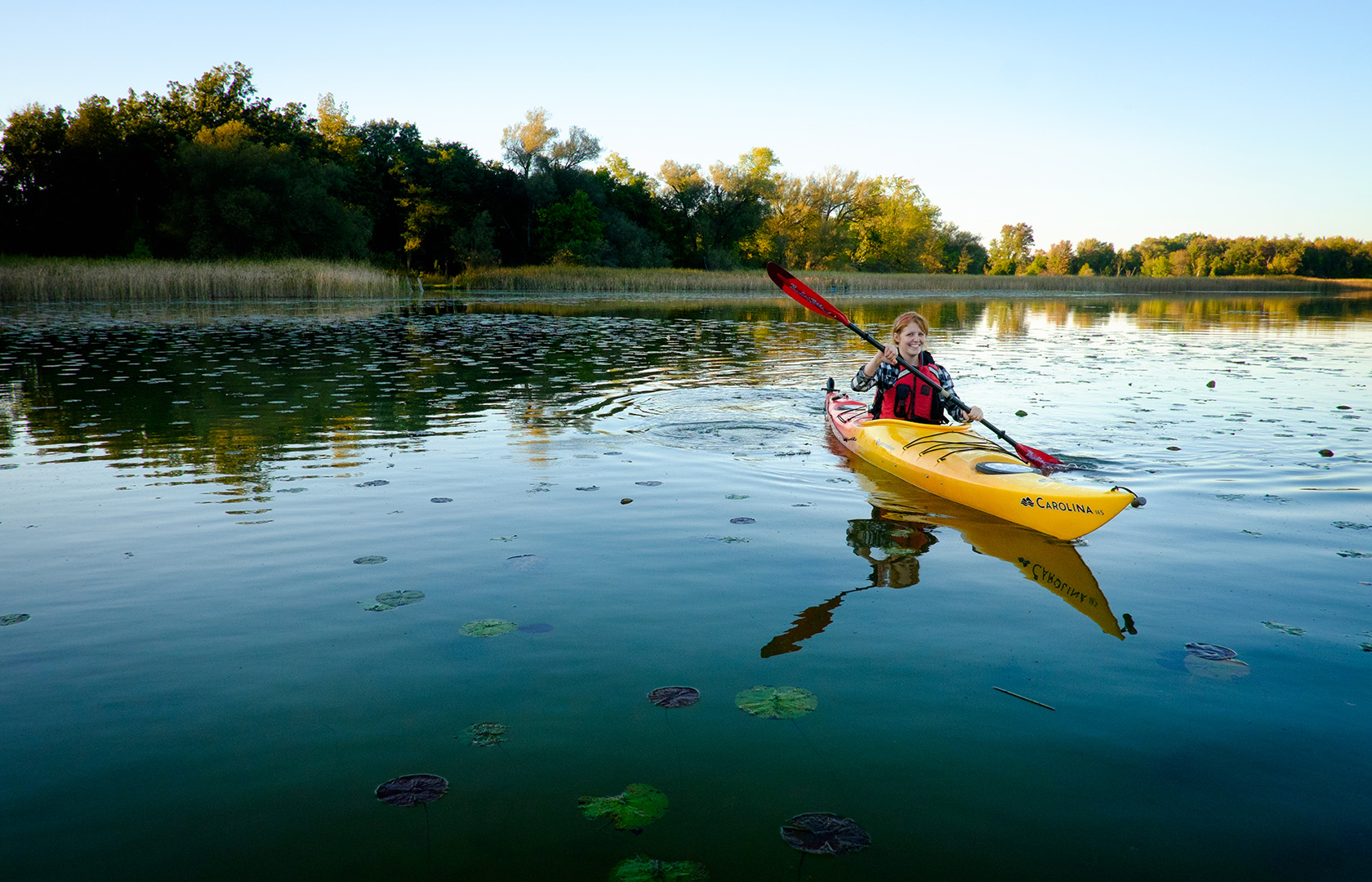 A person paddling a yellow kayak across a calm lake dotted with lily pads, surrounded by trees under a clear blue sky.