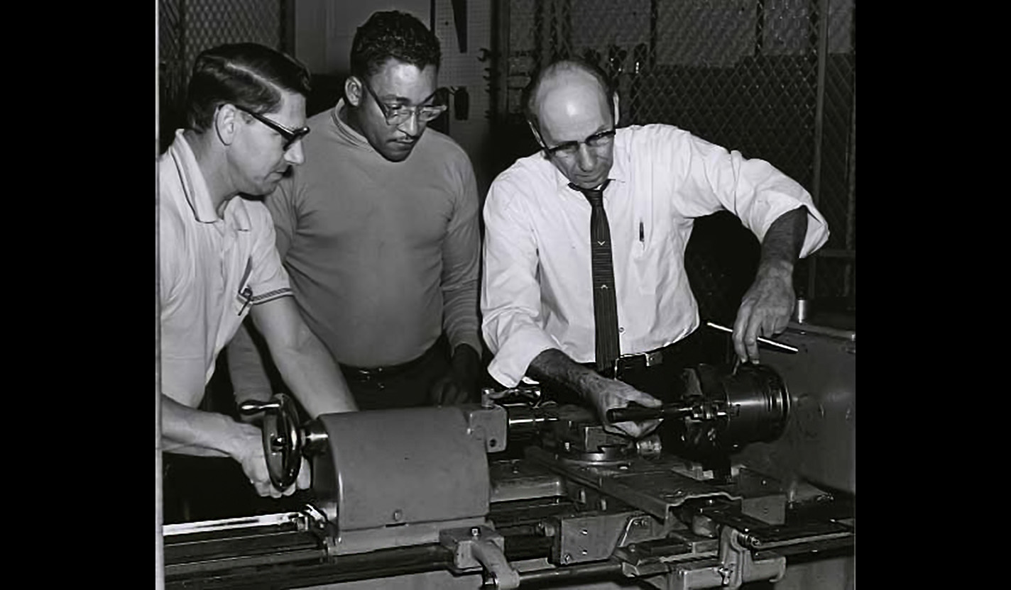 A black-and-white photo of two men watching an instructor use a metal lathe circa 1960.