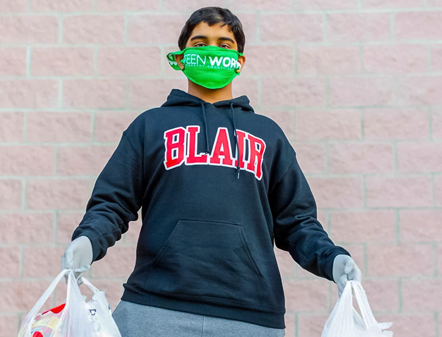 A young man in a black hoodie and green mask holds two grocery bags.