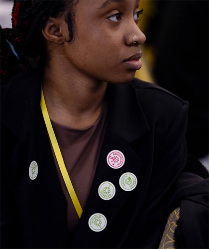 A young woman in a black blazer wears stickers that say: Resourceful, Growth Mindset, Optimism, Empathy, Persistence & Grit.