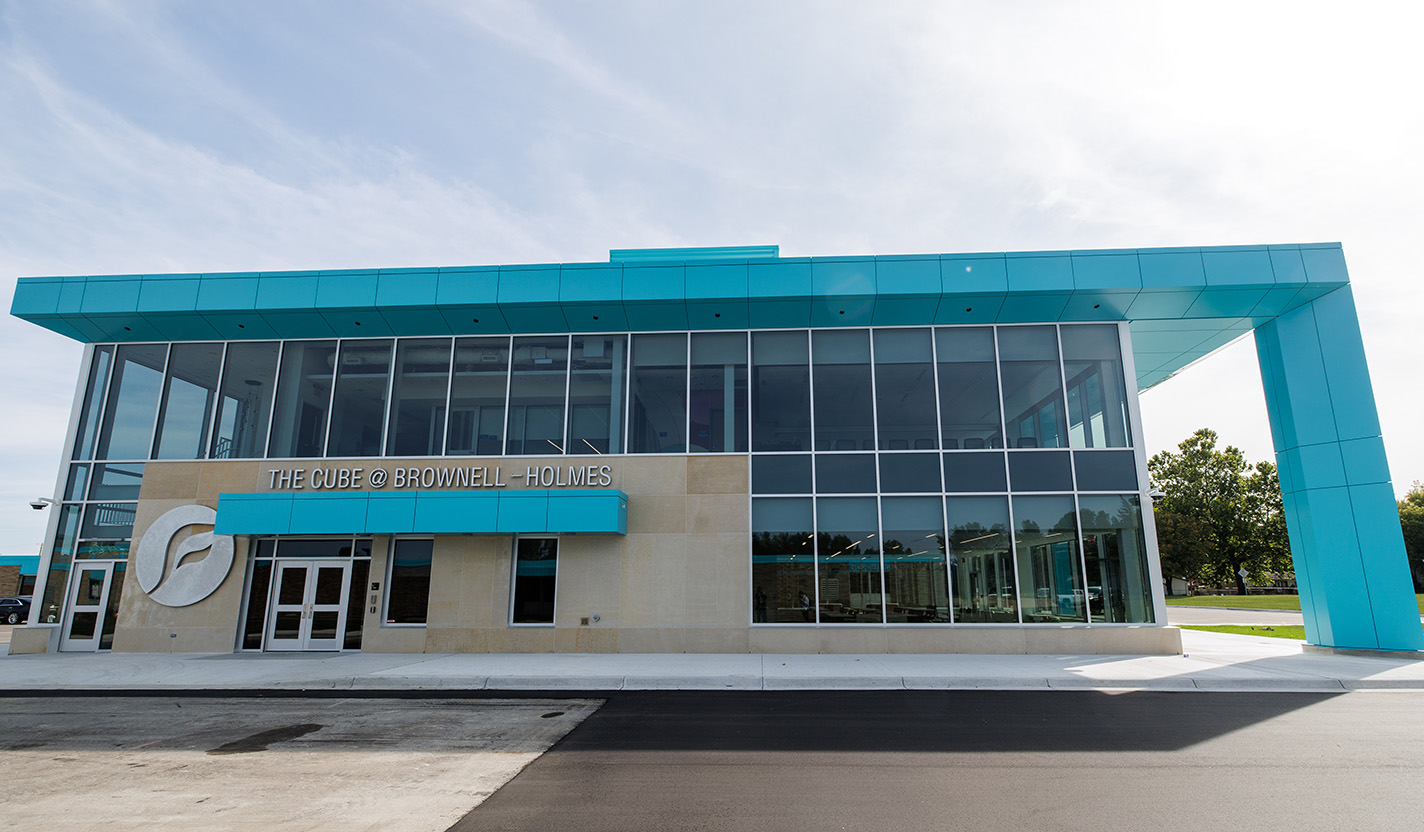 This image shows a a modern steel and glass school building in the shape of a cube with a light blue building frame and column, large windows and an asymetrical facade that is two stories high.
