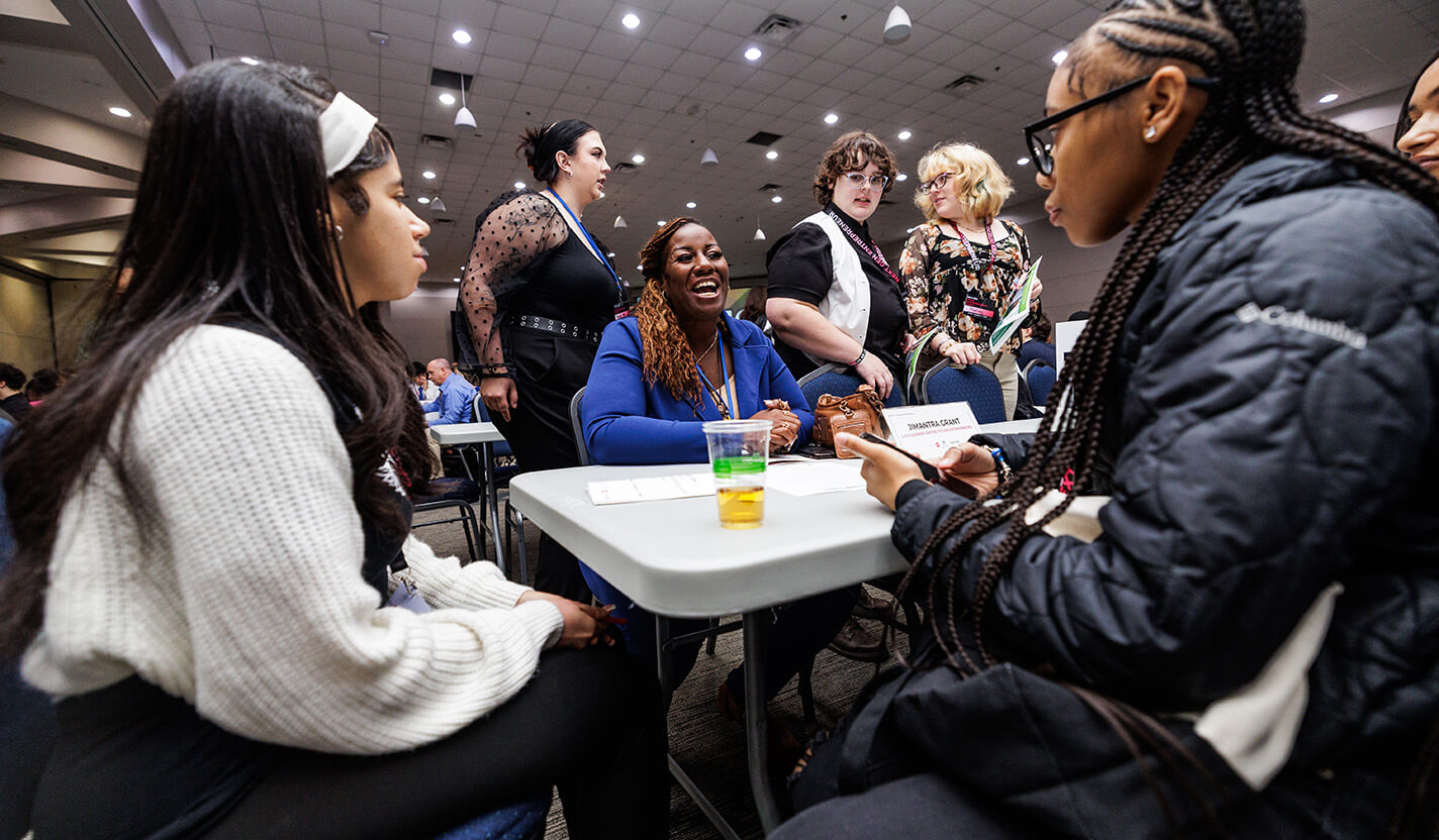 Woman in blue blazer laughs while speaking to two teenage girls at a table.