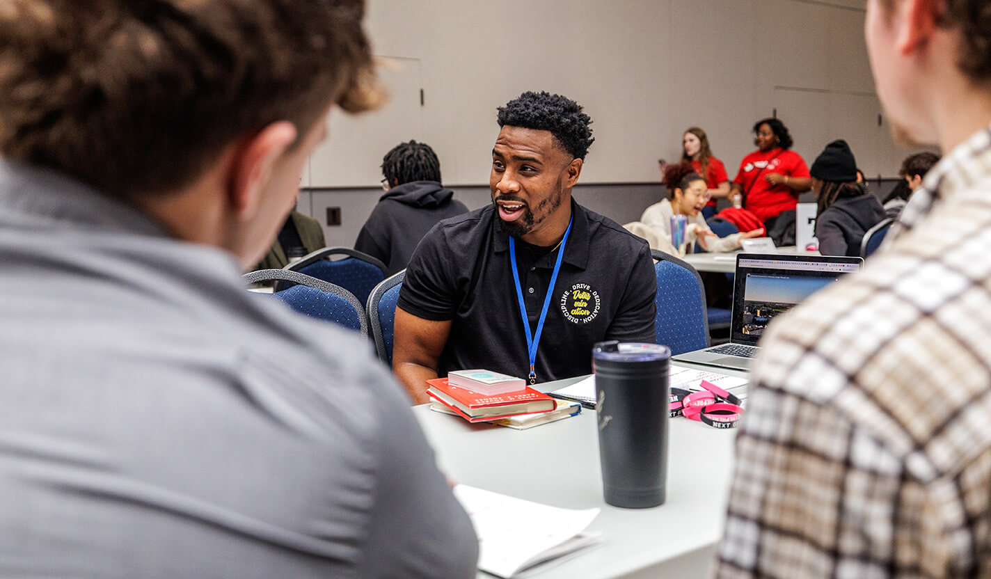 Man in black polo speaks with people at a table.
