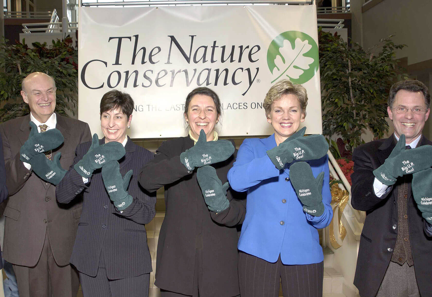 A group of smiling people stand in front of a Nature Conservancy banner holding up their hands in green mittens with the organization’s logo to form the shape of the state of Michigan.